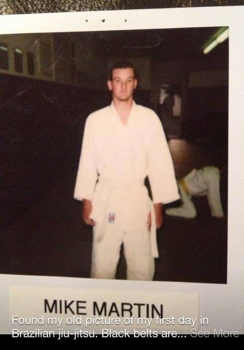 Mike Martin in a white Brazilian jiu-jitsu gi standing in a dojo with a fellow martial artist in the background.