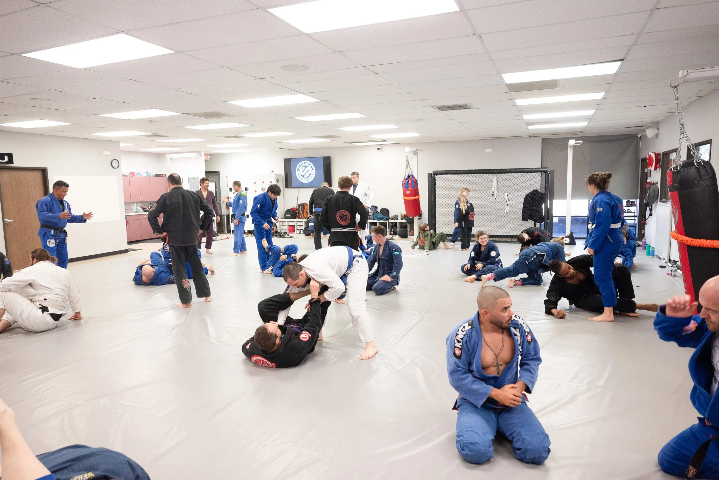 Martial arts class in a gym with students practicing ground fighting techniques. Participants wear gi uniforms, and some are engaged in grappling and rolling on a padded floor. The room has a high ceiling with bright lighting, hanging punching bags, and a cage. Several students and instructors are present, some observing and others instructing.