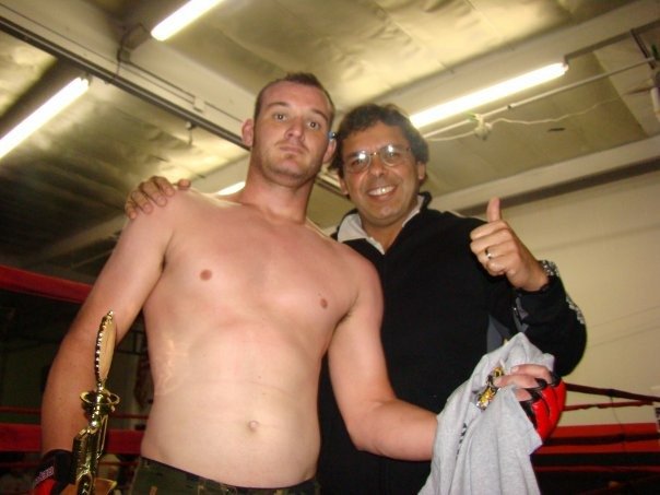 Joe Moreira and Mike standing together in a boxing ring, one shirtless and the other wearing glasses and a black jacket, with one giving a thumbs up.