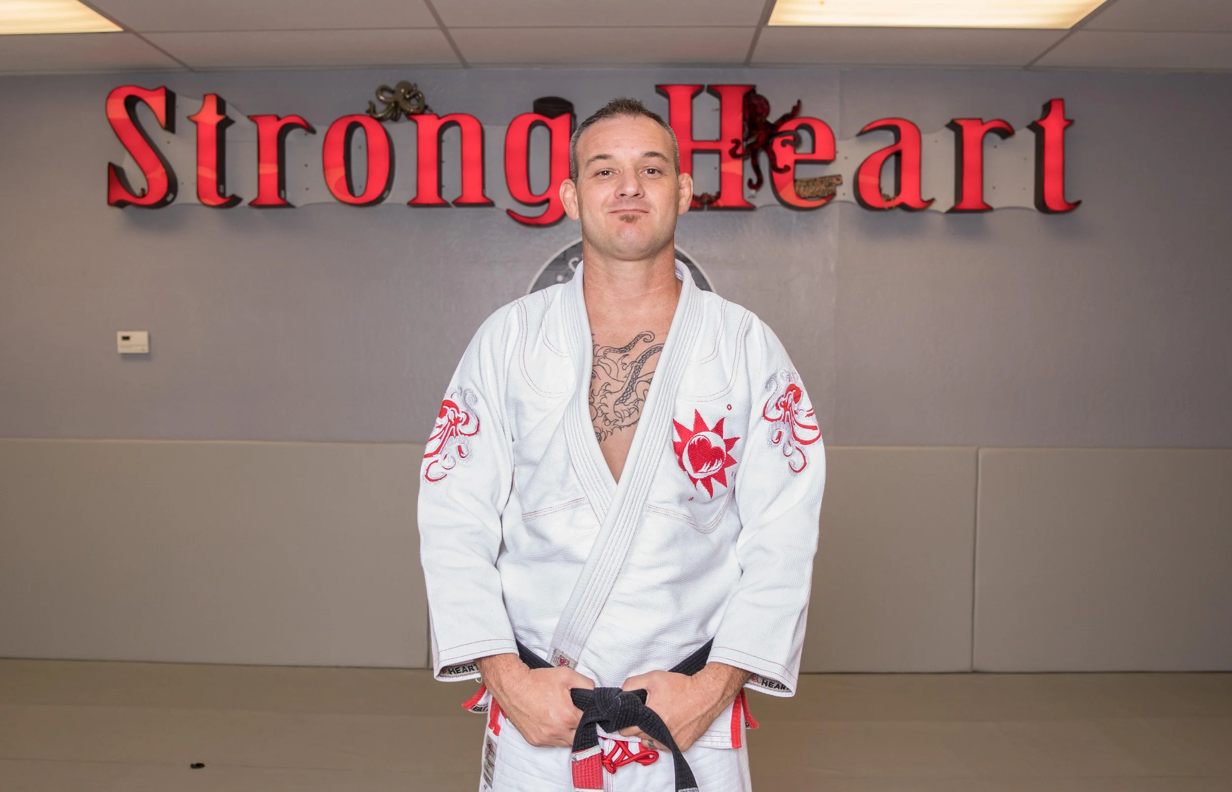 Mike in a white Brazilian Jiu-Jitsu gi standing in front of a wall with a red and black 'Strong Heart' sign. He has tattoos on his chest and is holding a black belt. The setting appears to be a martial arts studio or gym.
