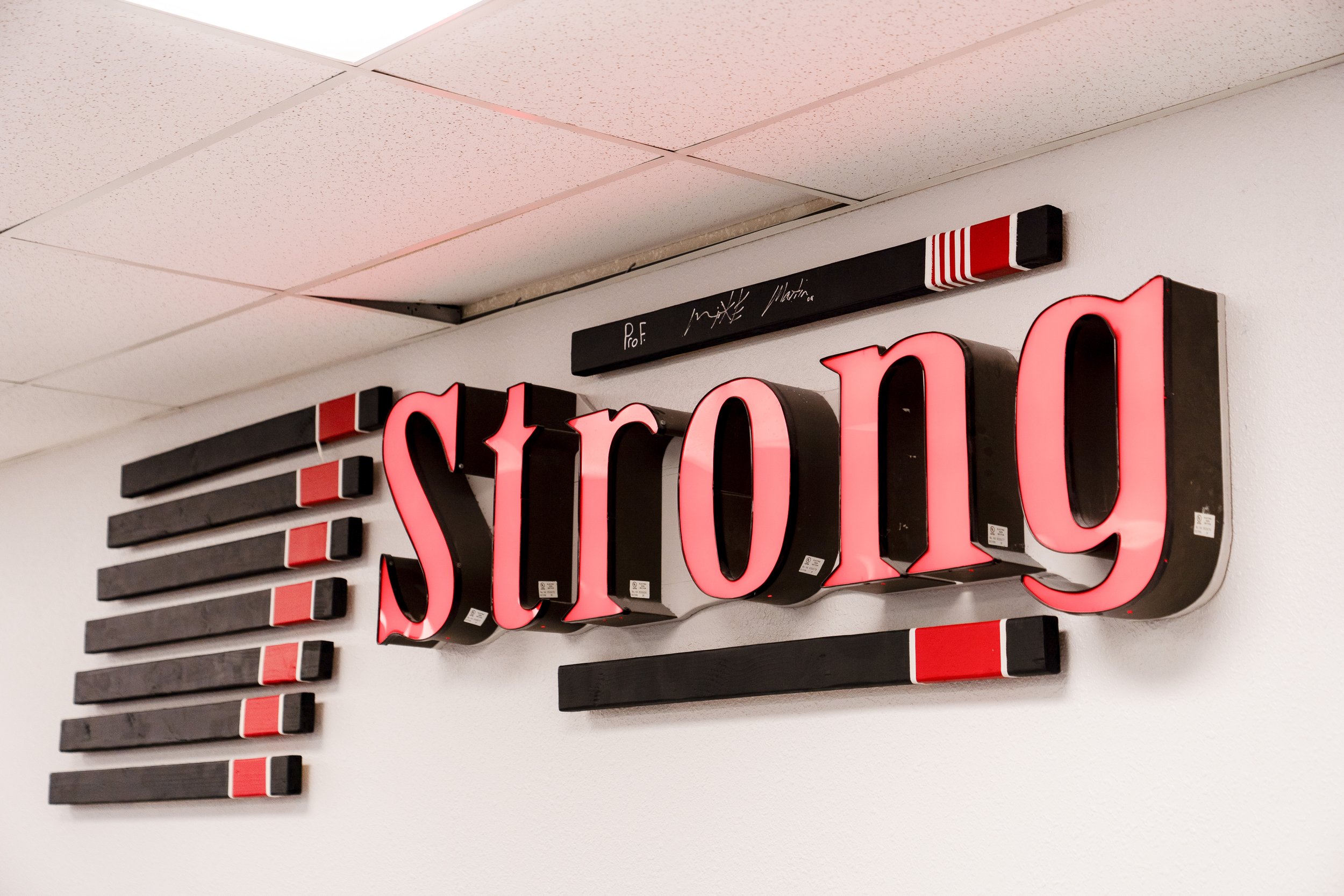 A large illuminated sign spelling out 'Strong' with black and red letters mounted on a wall, with a black metal bar above it and a black-and-white marker.  The sign is above BJJ Mats at the school.