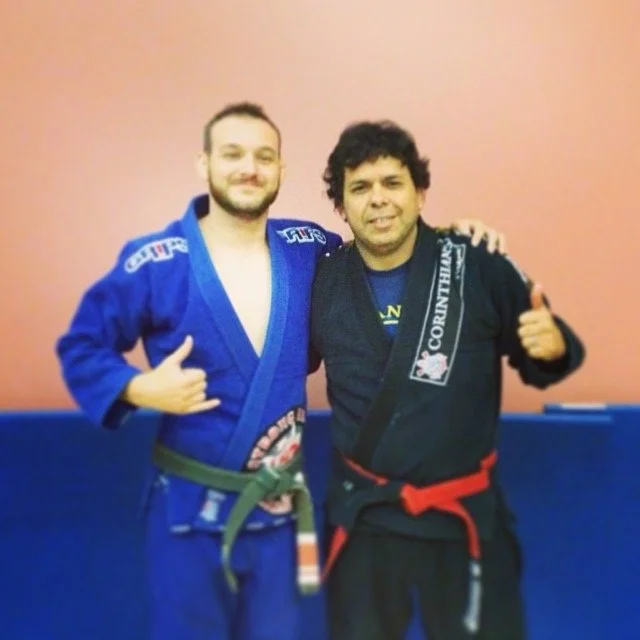 Joe Moreira and Mike in Brazilian Jiu-Jitsu gis smiling and posing together in a dojo, with one man wearing a blue gi and the other wearing a black gi, both showing thumbs up.
