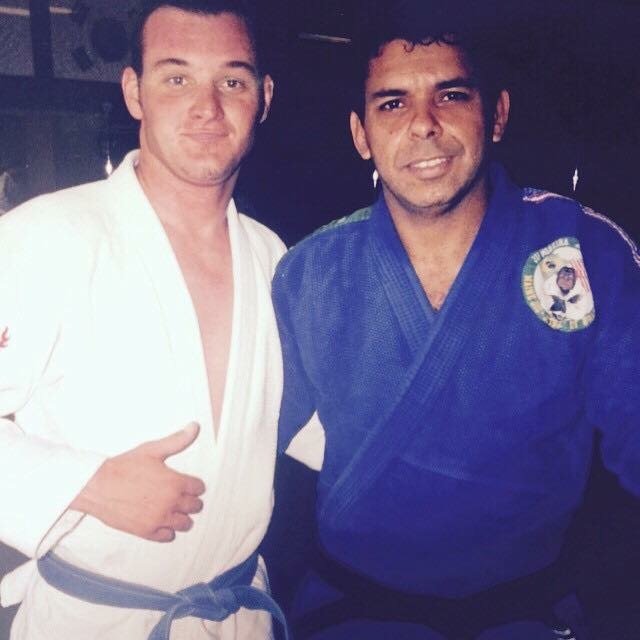 Joe Moreira and Mike in martial arts uniforms, one in a white gi, giving a thumbs-up, and the other in a blue gi with a badge, posing together.