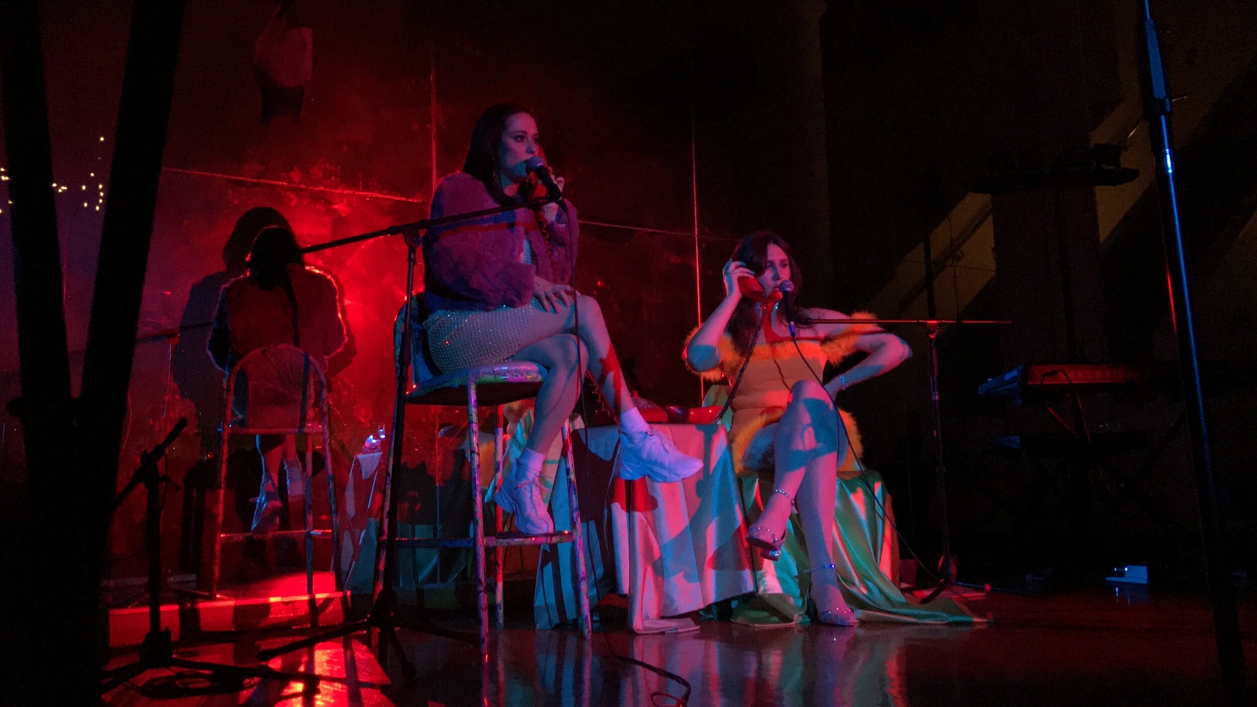 Two women sit on a stage singing while bathed in red and blue light.