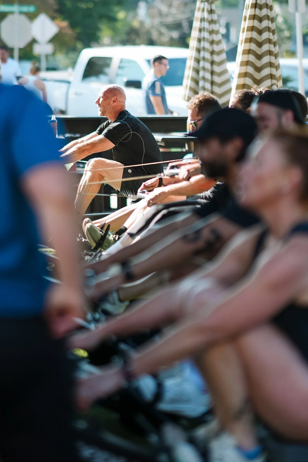People participating in an outdoor fitness class, seated in a row on exercise bikes under large striped umbrellas on a sunny day.
