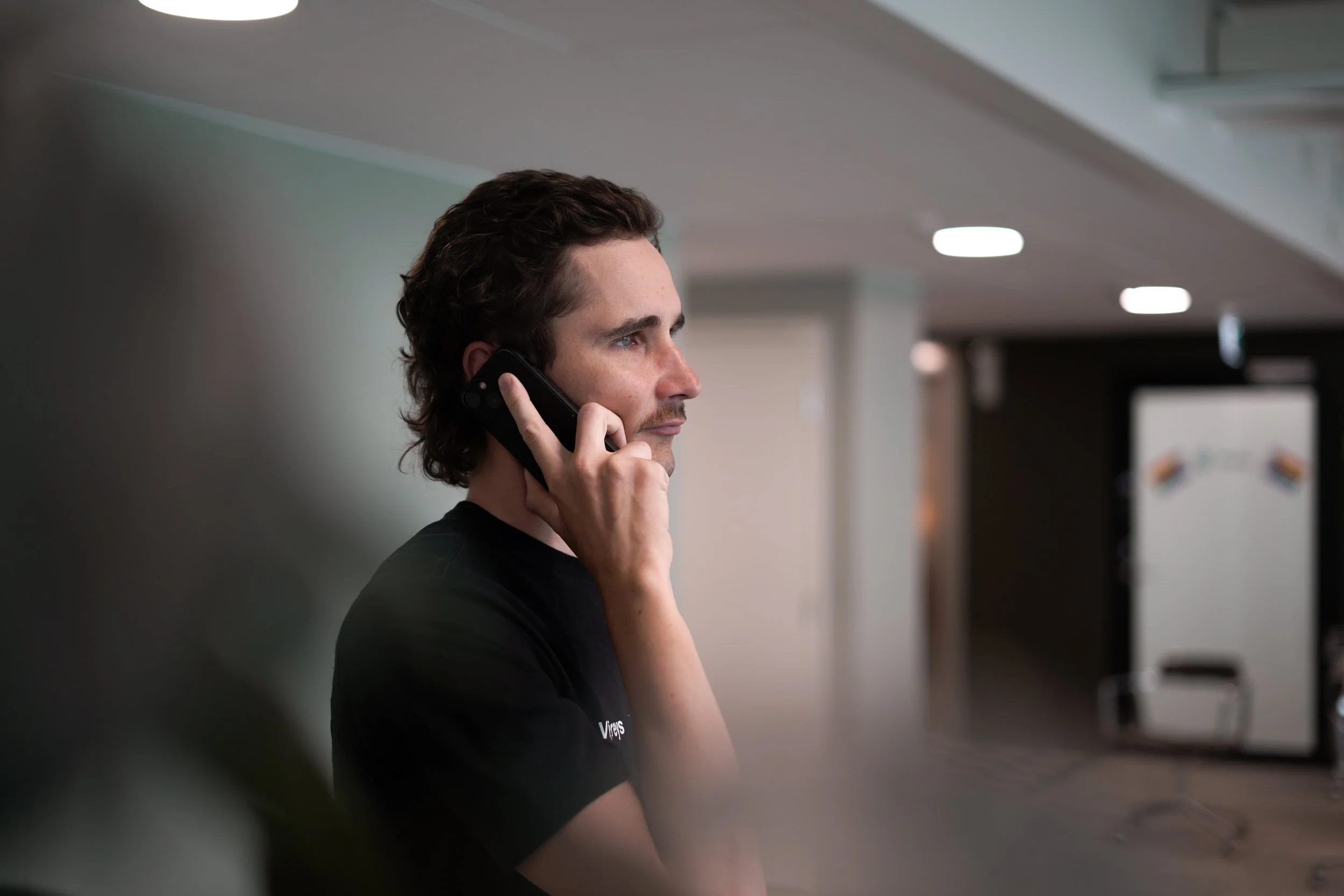 Nuori mies puhuttaa puhelimessa sisätiloissa, taustalla vierashuone tai vastaanottoalue.