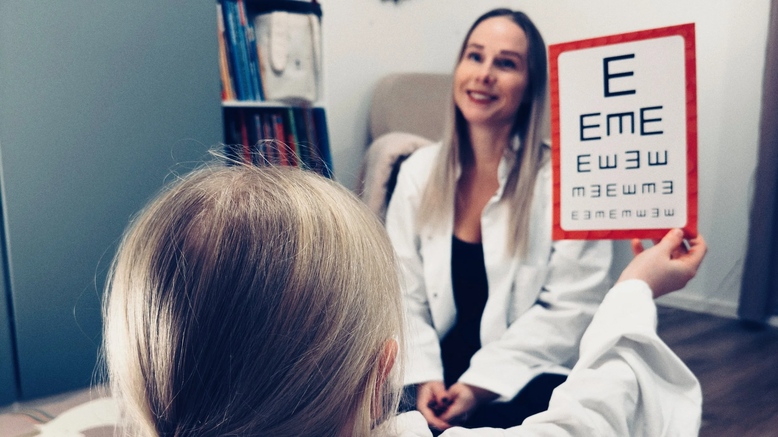 Kuvassa pieni lapsi leikkii lääkäriä ja pitää näkötestiä kädessään. Taustalla lapsen äiti leikkii potilasta.