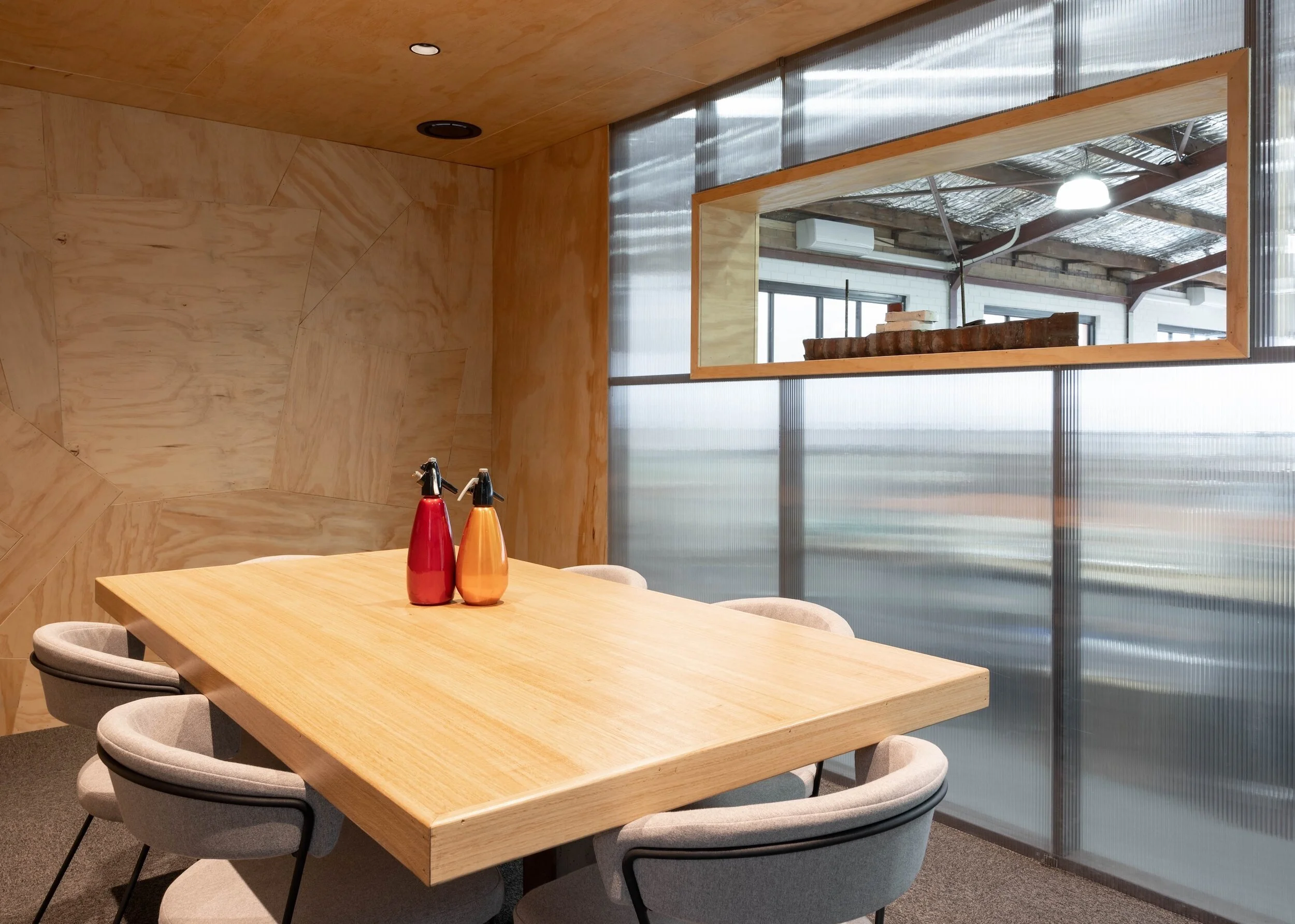 A wooden dining table with six cushioned chairs in a modern interior space, with a large window and a wooden shelf behind it.