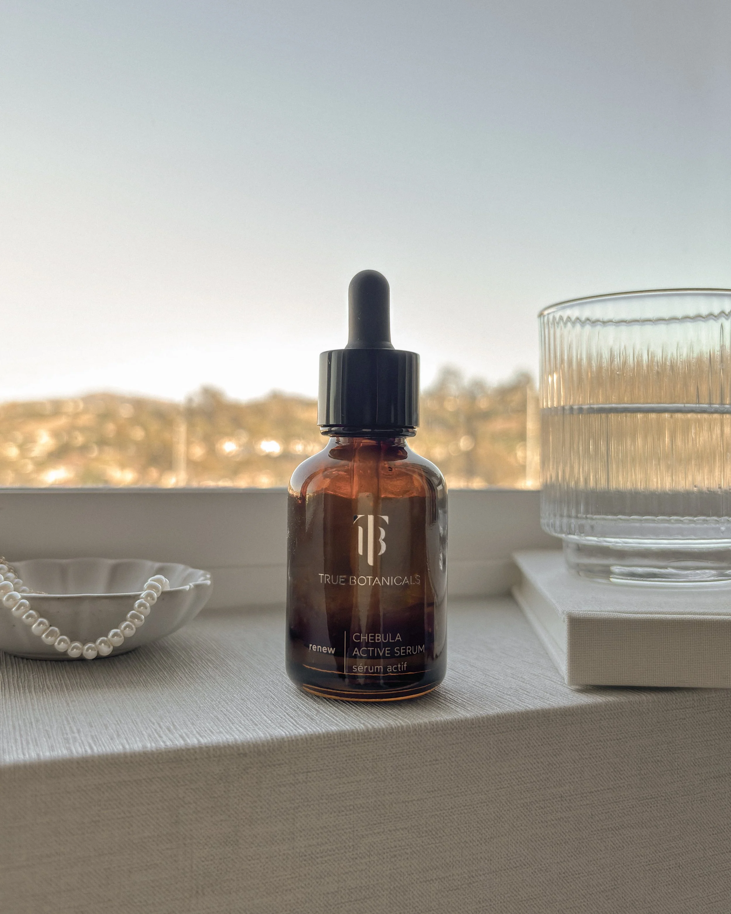 True Botanicals Chebula Active Serum on a table by a window with a pearl dish and glass of water.