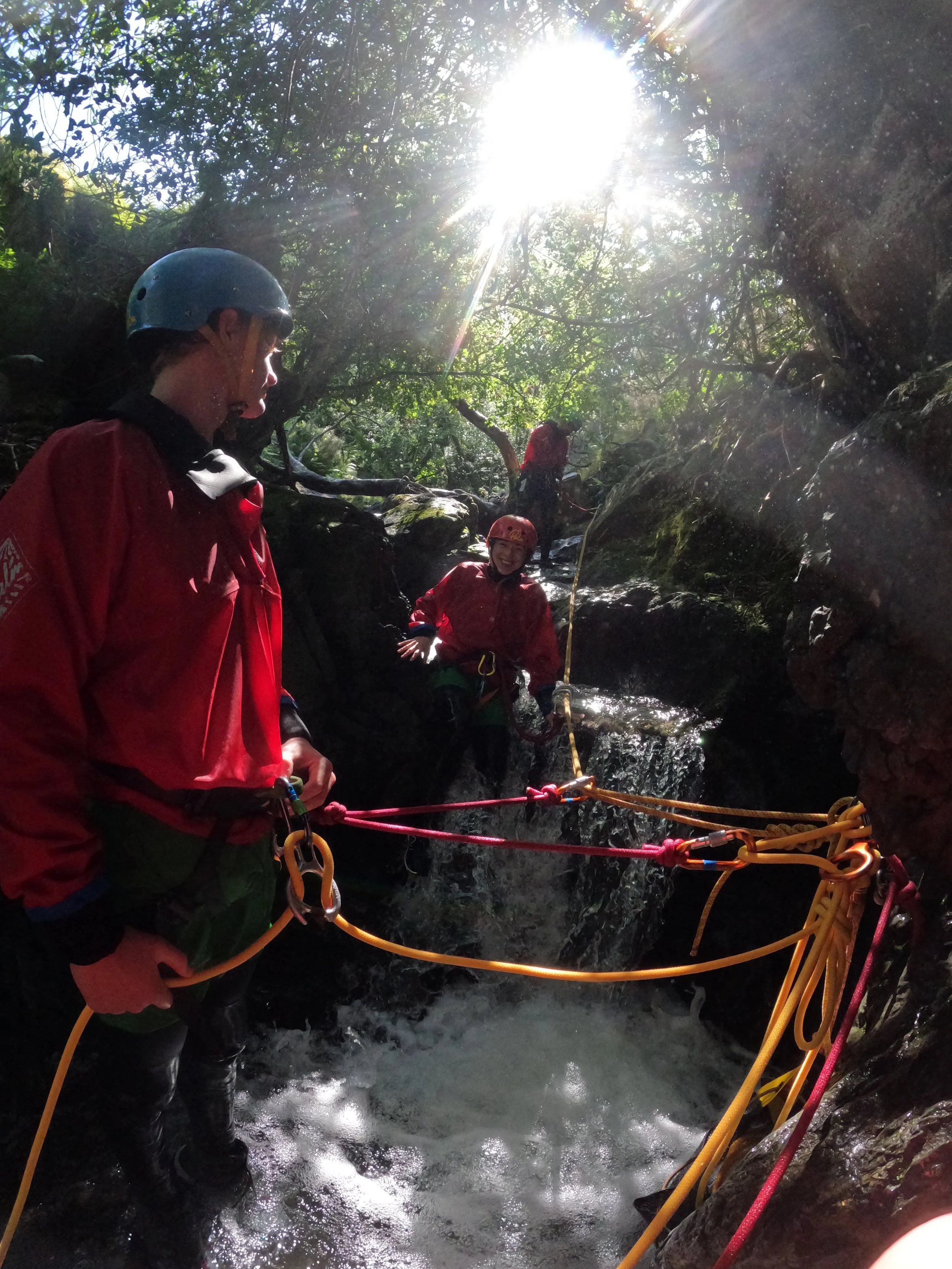 Three people wearing helmets and red jackets canyoning in a rocky creek, with sunlight shining through the trees above.