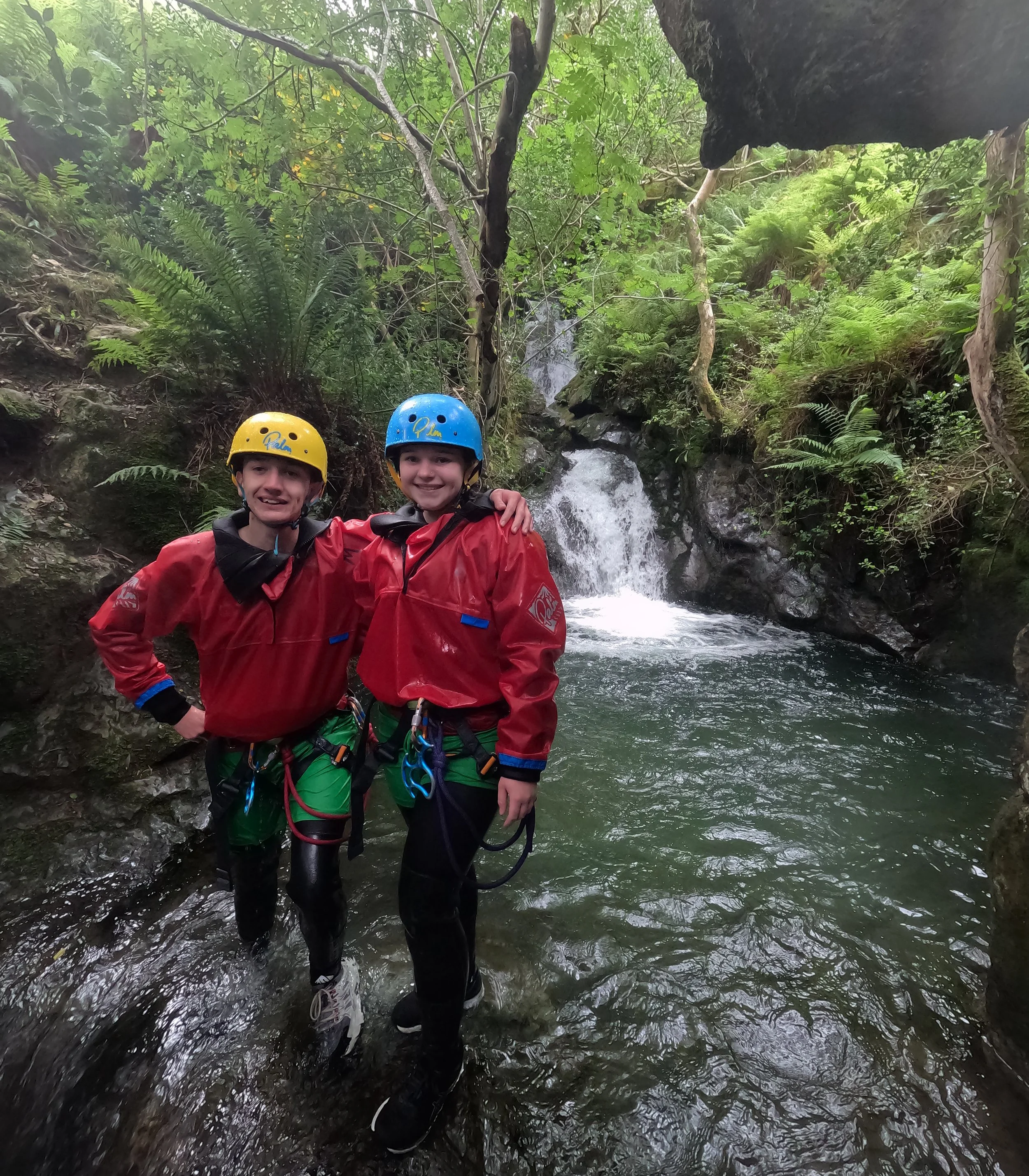 Two people wearing red jackets and helmets with climbing gear standing in a stream, surrounded by lush green forest and rocks, with a small waterfall behind them.