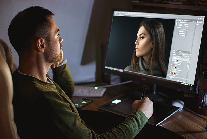 A retoucher using a pen tablet to hand retouch a woman's portrait on a computer monitor in photo editing software.
