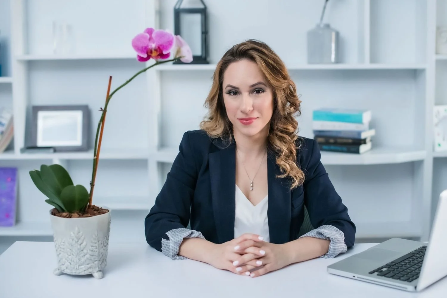 Professional woman in a navy blazer seated at a white office desk with a laptop and pink orchid, branding photoshoot portrait
