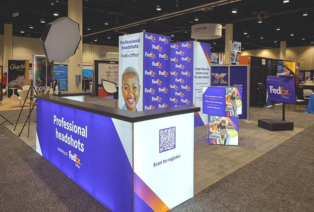 Branded professional headshot booth at a corporate trade show, with a sign-up counter displaying a scan-to-register QR code and sponsor graphics