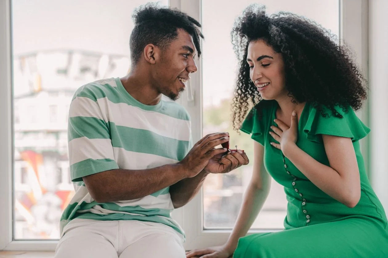 Man presenting an engagement ring to a smiling woman by a window, the moment an engagement photo session usually follows