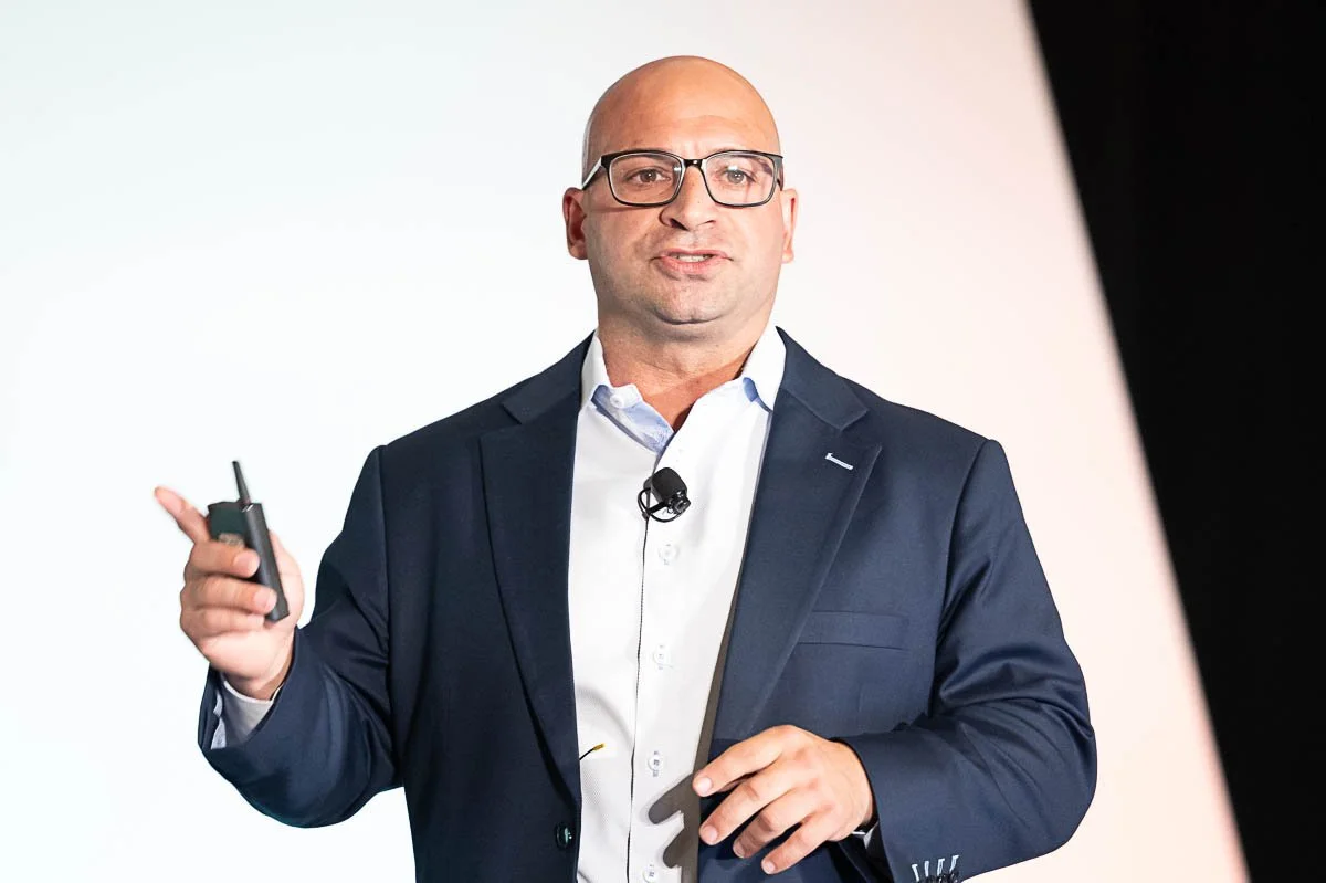 Corporate keynote speaker in a navy suit and glasses holding a presentation remote on a brightly lit conference stage