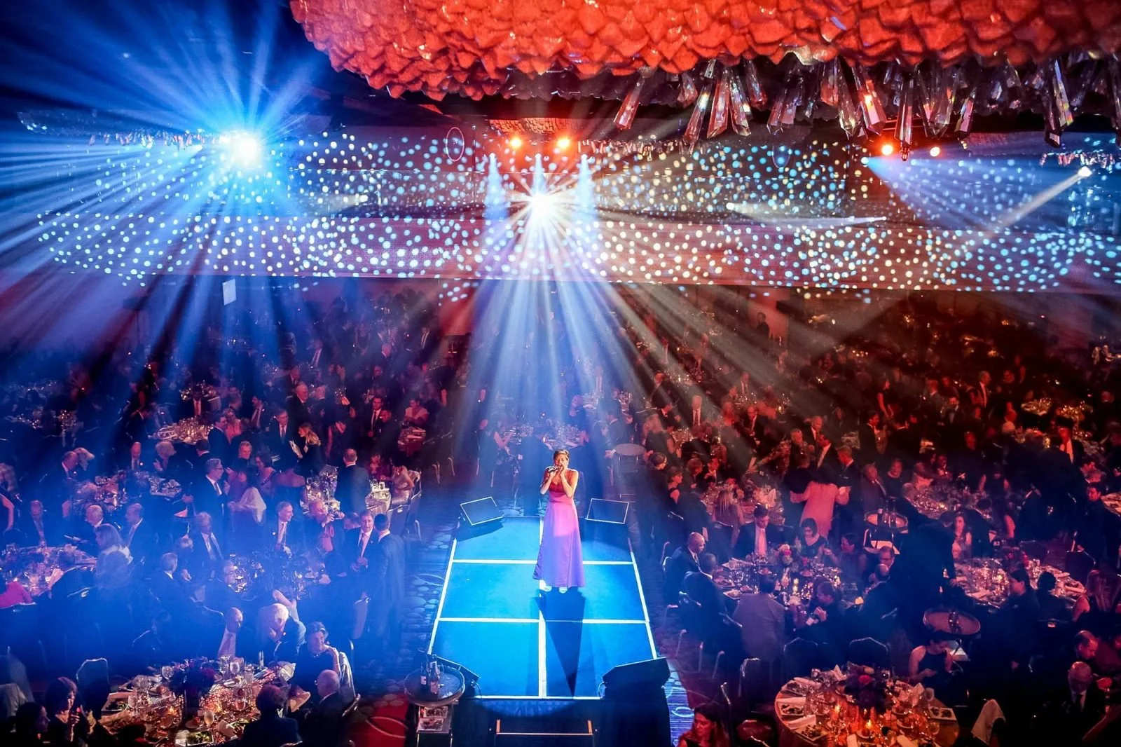 Singer in a pink gown performing on a blue-lit runway stage at a large corporate gala, with guests seated at round tables under dramatic red and blue stage lighting.