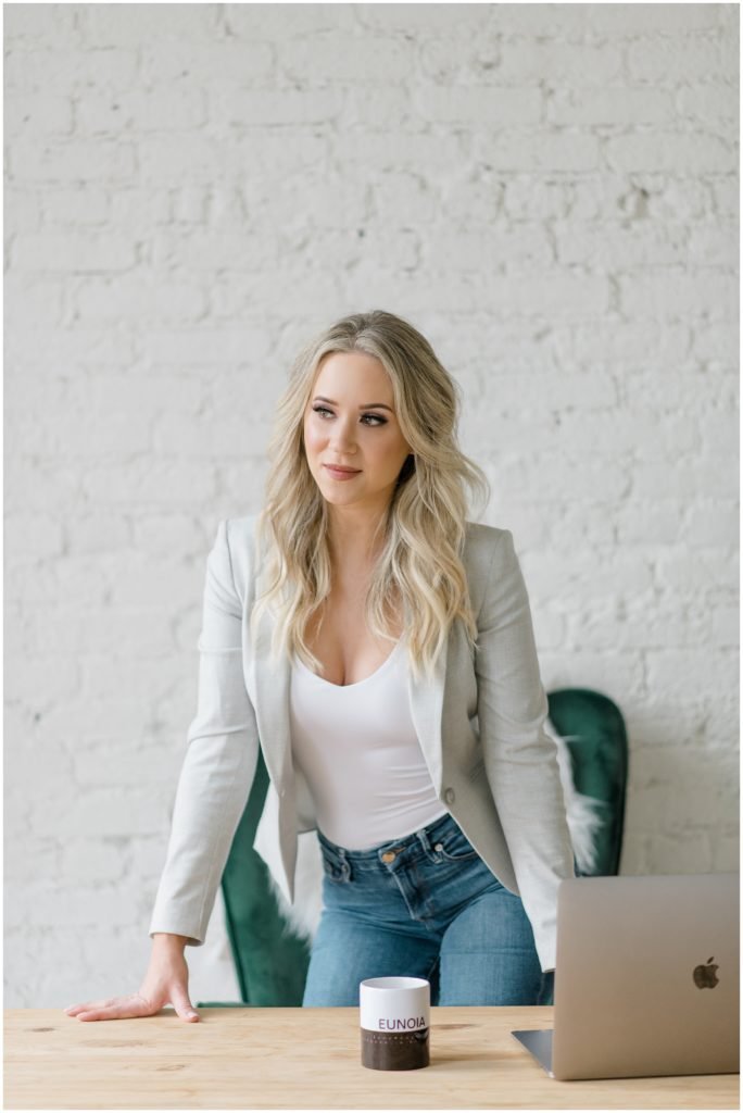Blonde entrepreneur in a light gray blazer and jeans standing at a wooden table with a laptop and coffee mug, personal branding session