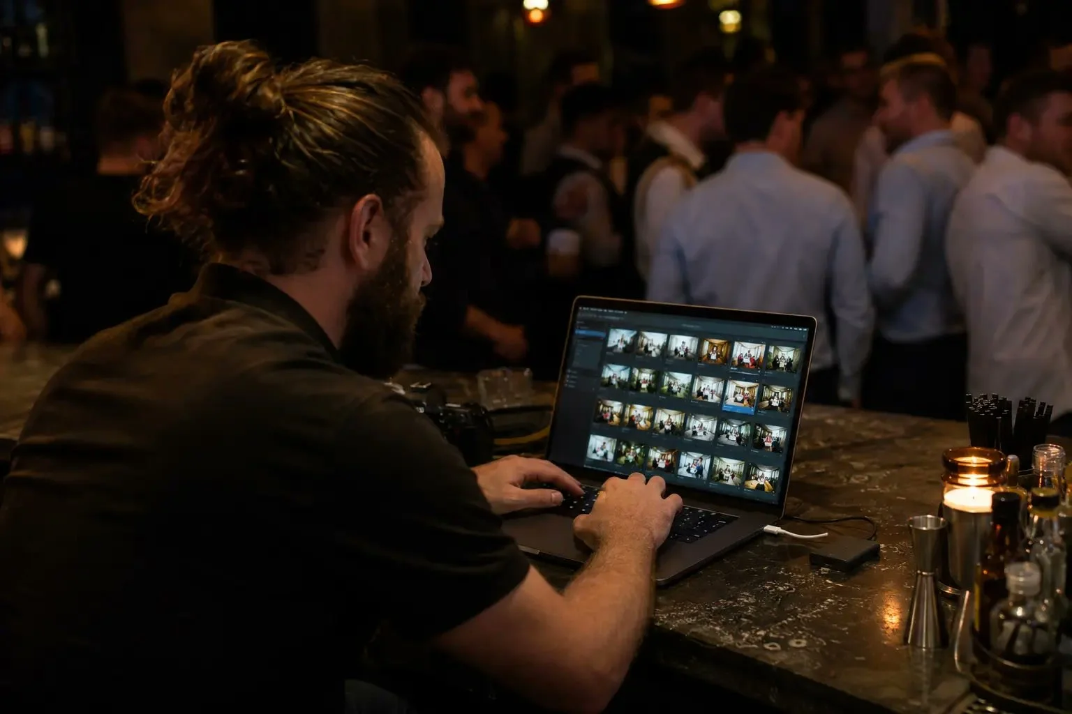 Event photographer editing and uploading photos on a laptop at a venue bar while guests mingle behind him