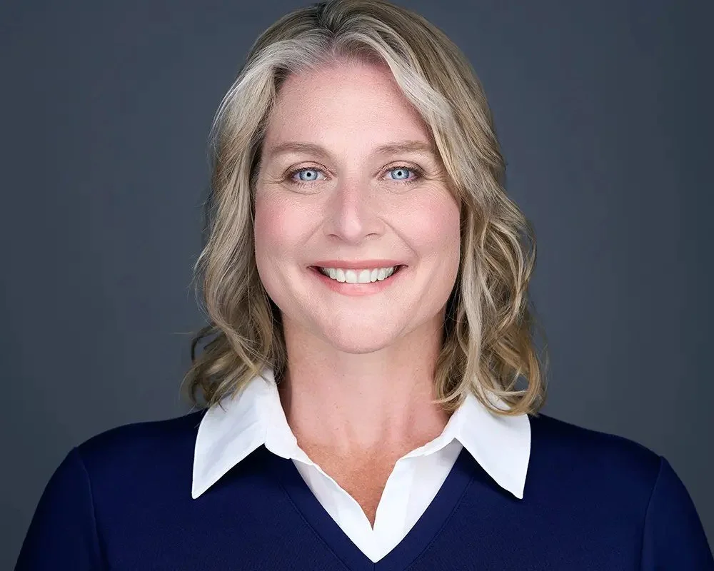 Smiling professional woman with blonde hair wearing a navy sweater over a white collared shirt in a studio headshot against a gray background