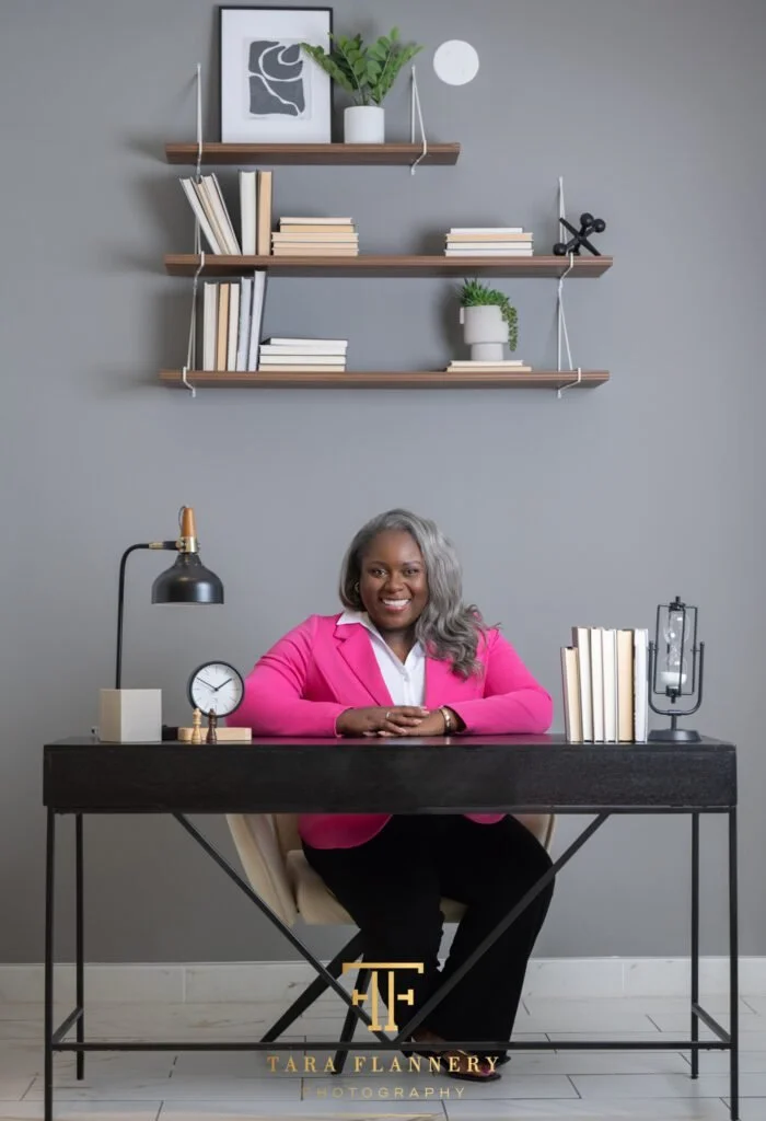 Smiling businesswoman in a bright pink blazer seated at a styled dark desk with books, personal branding portrait