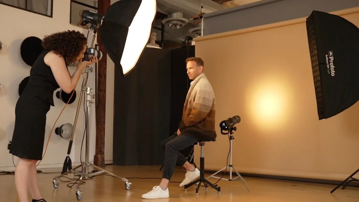 Behind the scenes of a professional headshot session: a photographer shooting a seated man in a studio with a large softbox key light, a Profoto strip light, and a beige seamless backdrop.
