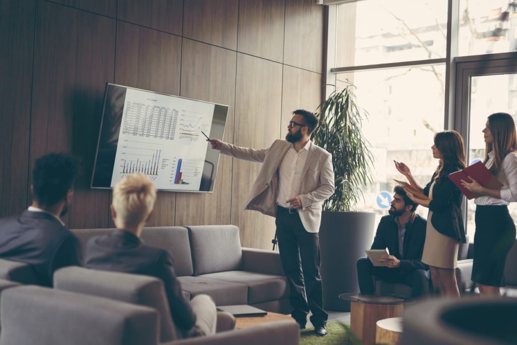 Business team in a planning meeting with a man pointing at charts on a wall-mounted screen while colleagues listen.