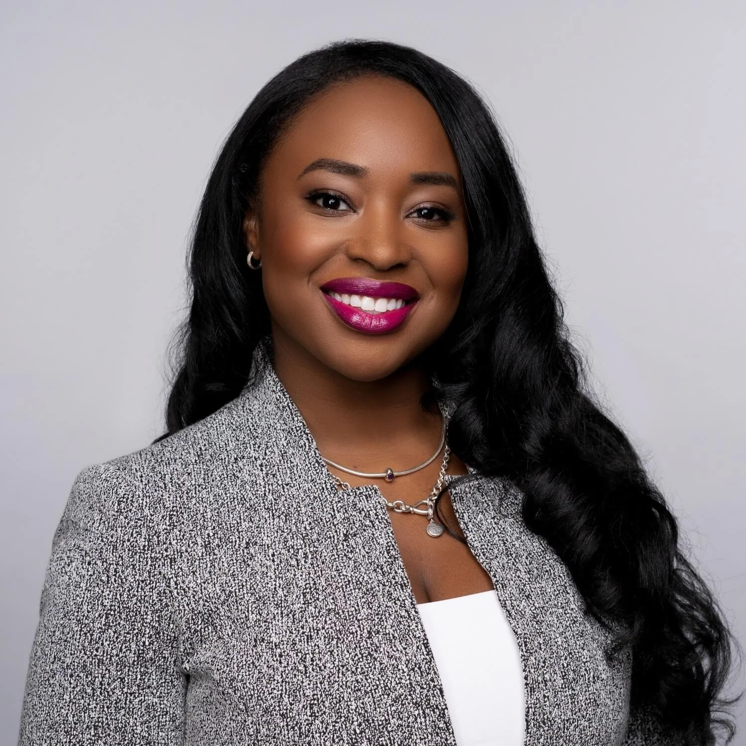 Professional headshot of a smiling woman with long dark hair wearing a gray blazer against a plain light gray studio background