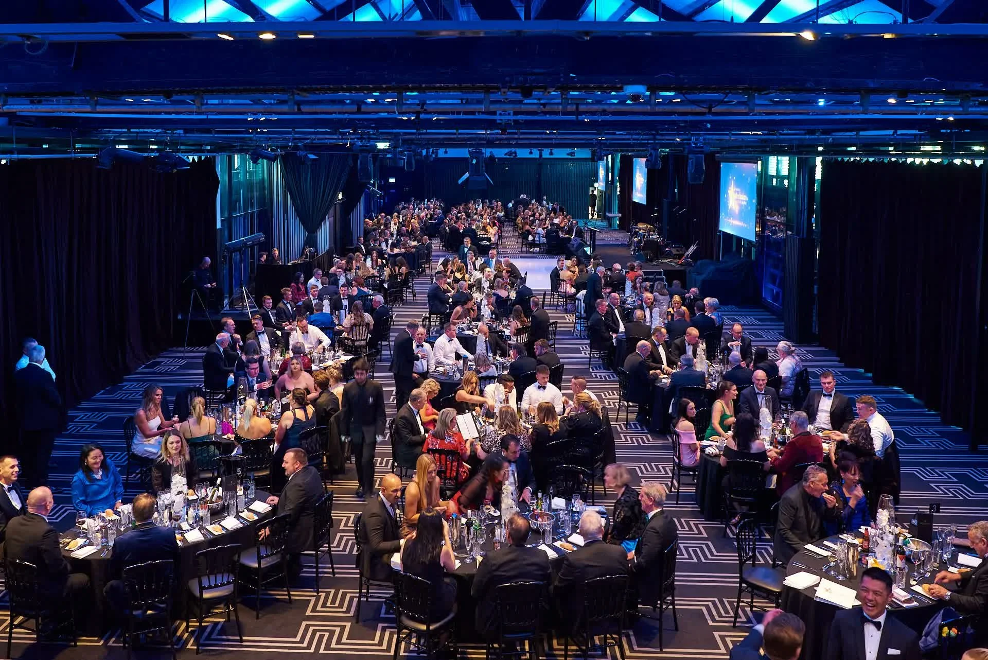 Wide view of a large black tie corporate gala dinner with hundreds of guests seated at round tables under blue stage lighting