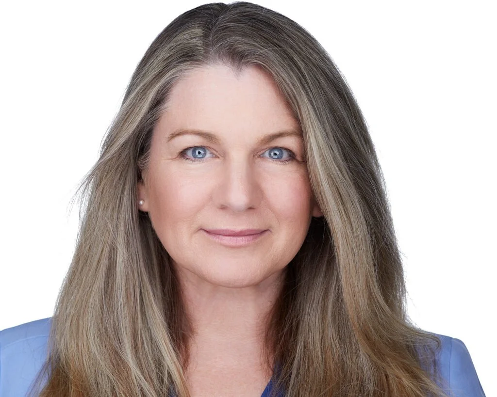 Close-up studio headshot of a woman with long blonde hair and blue eyes against a white background