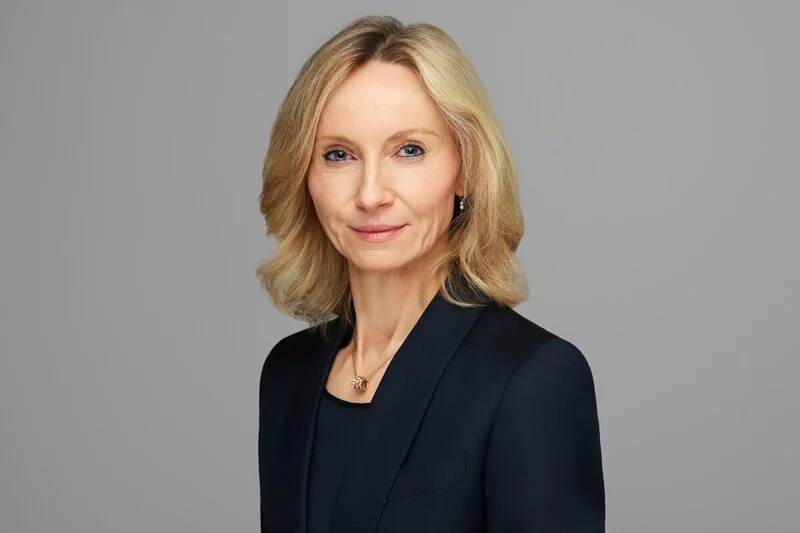 Professional headshot of a smiling blonde woman wearing a dark navy blazer and a delicate pendant necklace against a light gray studio background