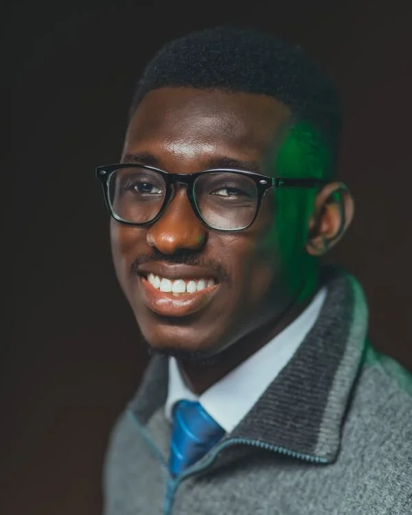 Smiling professional headshot of a man with glasses wearing a gray blazer and blue tie against a dark background