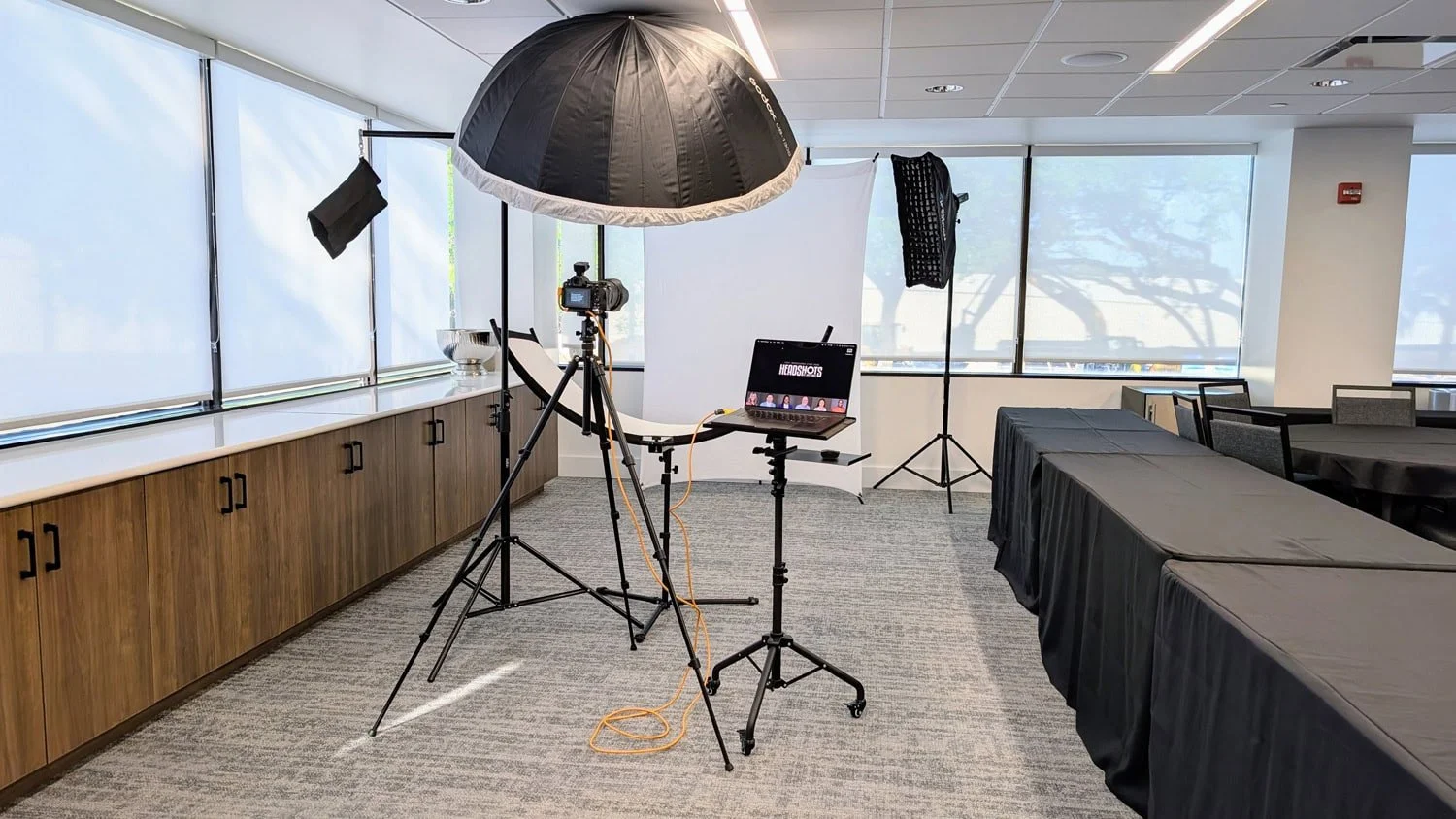 Behind-the-scenes view of an on-site headshot station in a conference room, with studio strobes and a large softbox, seamless backdrop, camera on tripod, and laptop showing tethered preview