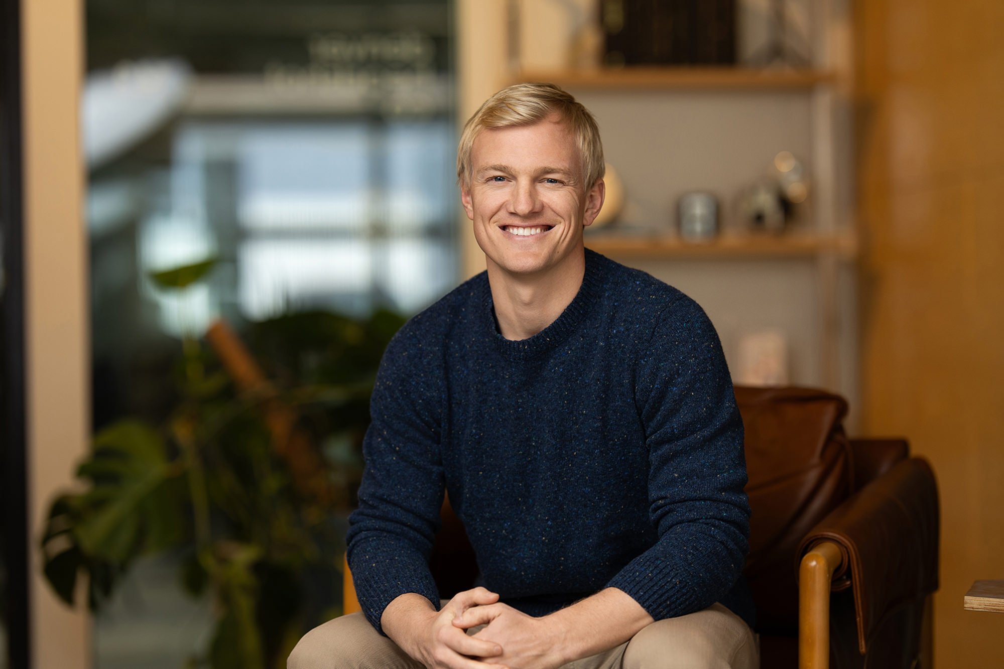 Smiling blond man in a navy sweater seated in a leather chair during an office headshot session