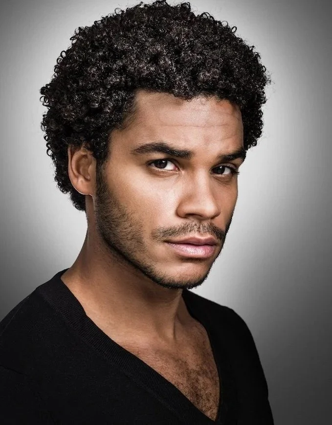 Los Angeles actor headshot of a man with dark curly hair and light stubble in a black v-neck shirt against a gray studio backdrop