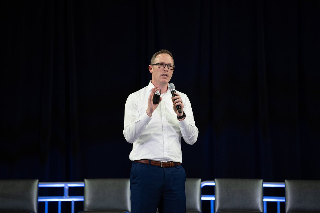 Keynote speaker in a white shirt holding a microphone on a dark conference stage with panel chairs behind him.