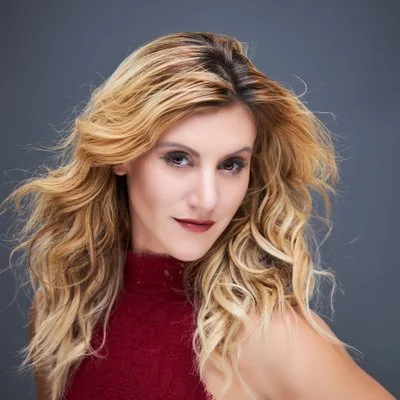 Studio headshot of a blonde woman with long wavy hair wearing a burgundy lace top against a gray studio background