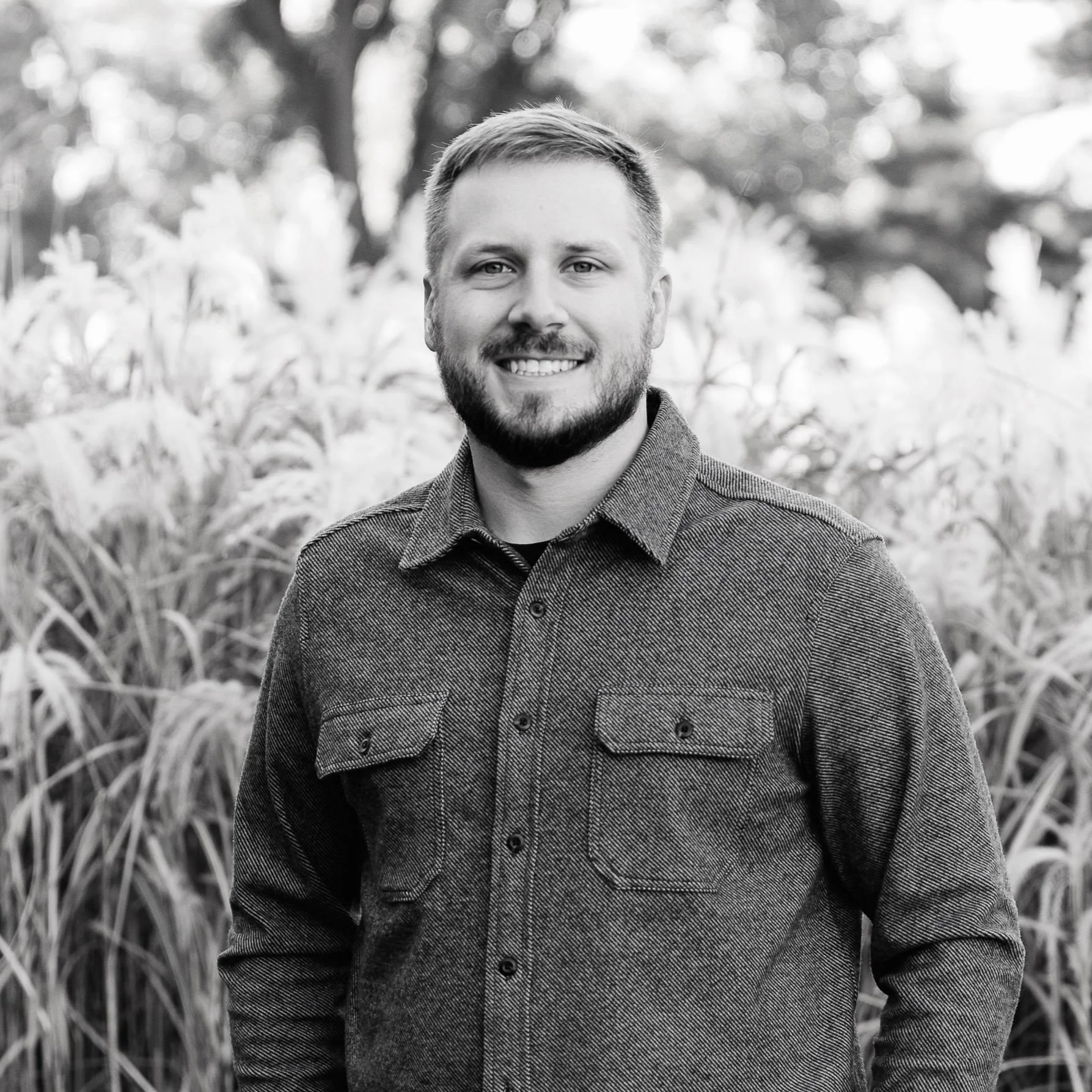 A man with short hair and a beard smiling outdoors, standing in front of tall grasses or plants, in black and white.
