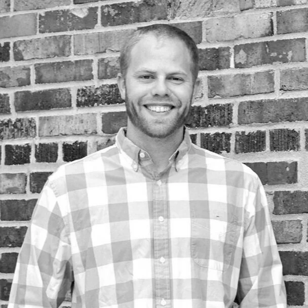 A smiling man with short hair and a beard, wearing a checkered button-up shirt, standing in front of a brick wall.