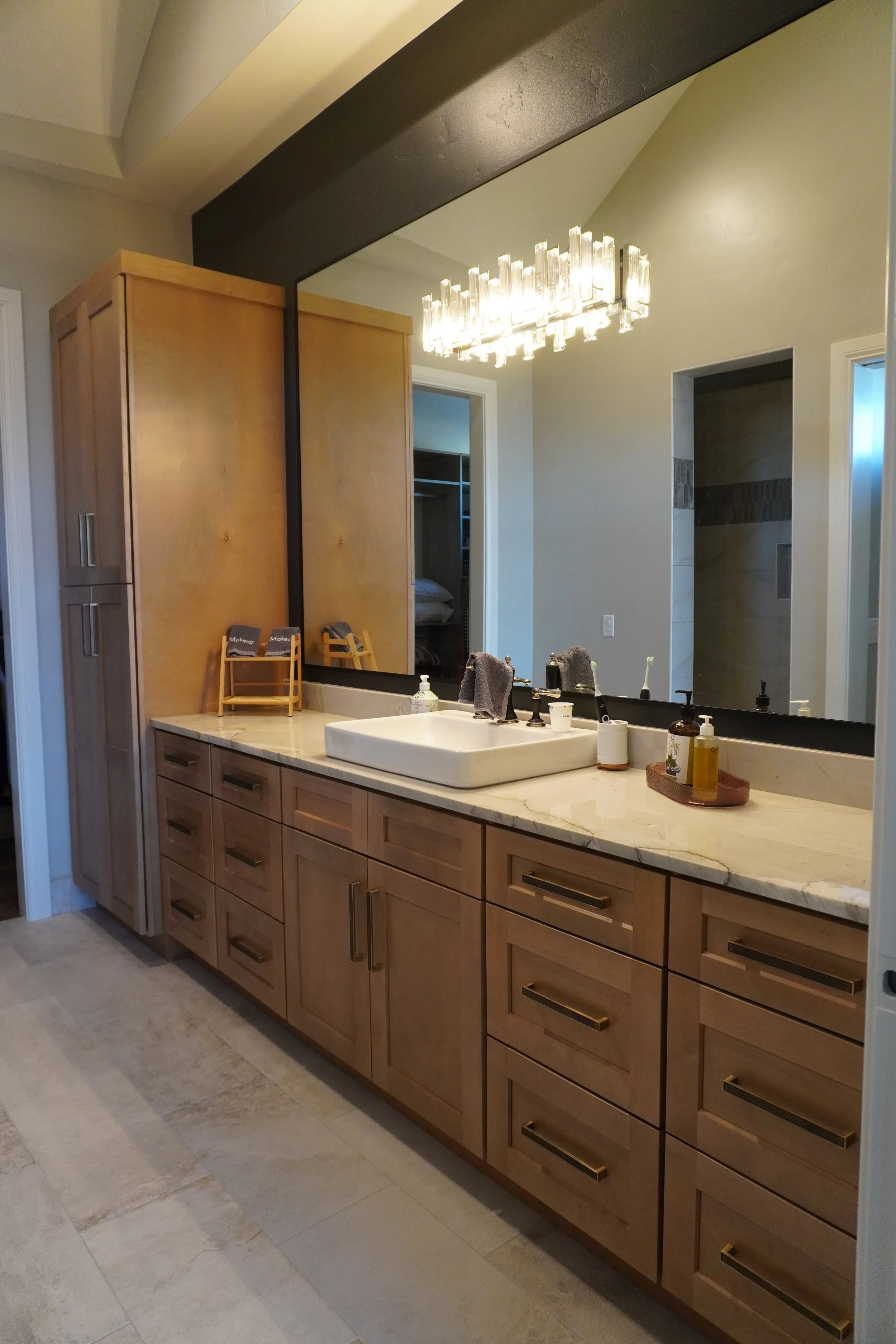 Modern bathroom vanity with wooden cabinets, marble countertop, rectangular vessel sink, large mirror, and modern chandelier light fixture.