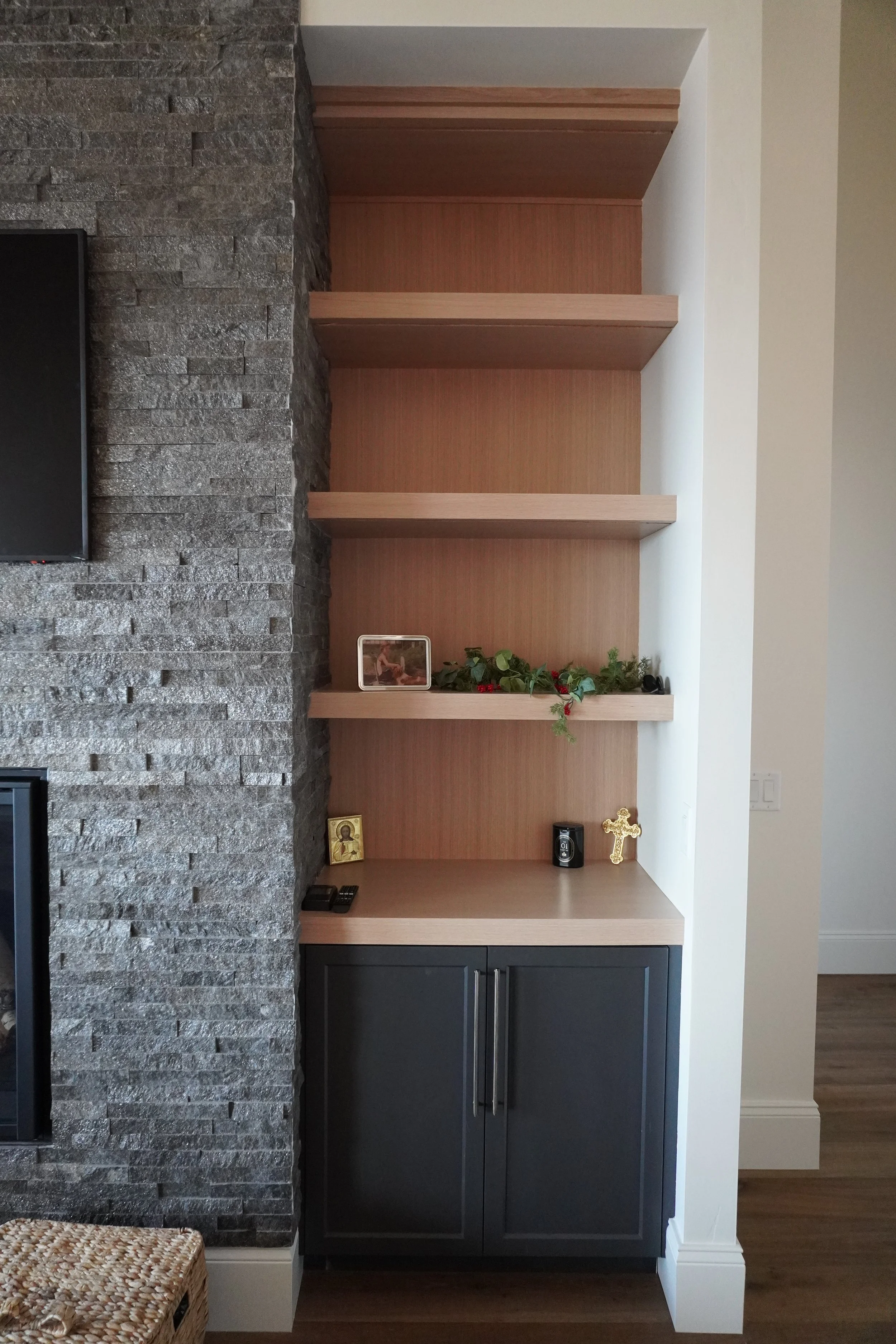 Modern built-in shelving unit with three open shelves, decorated with a small framed picture, greenery, candles, religious icons, and a golden cross, below a dark cabinet with silver handles, next to a stone wall.