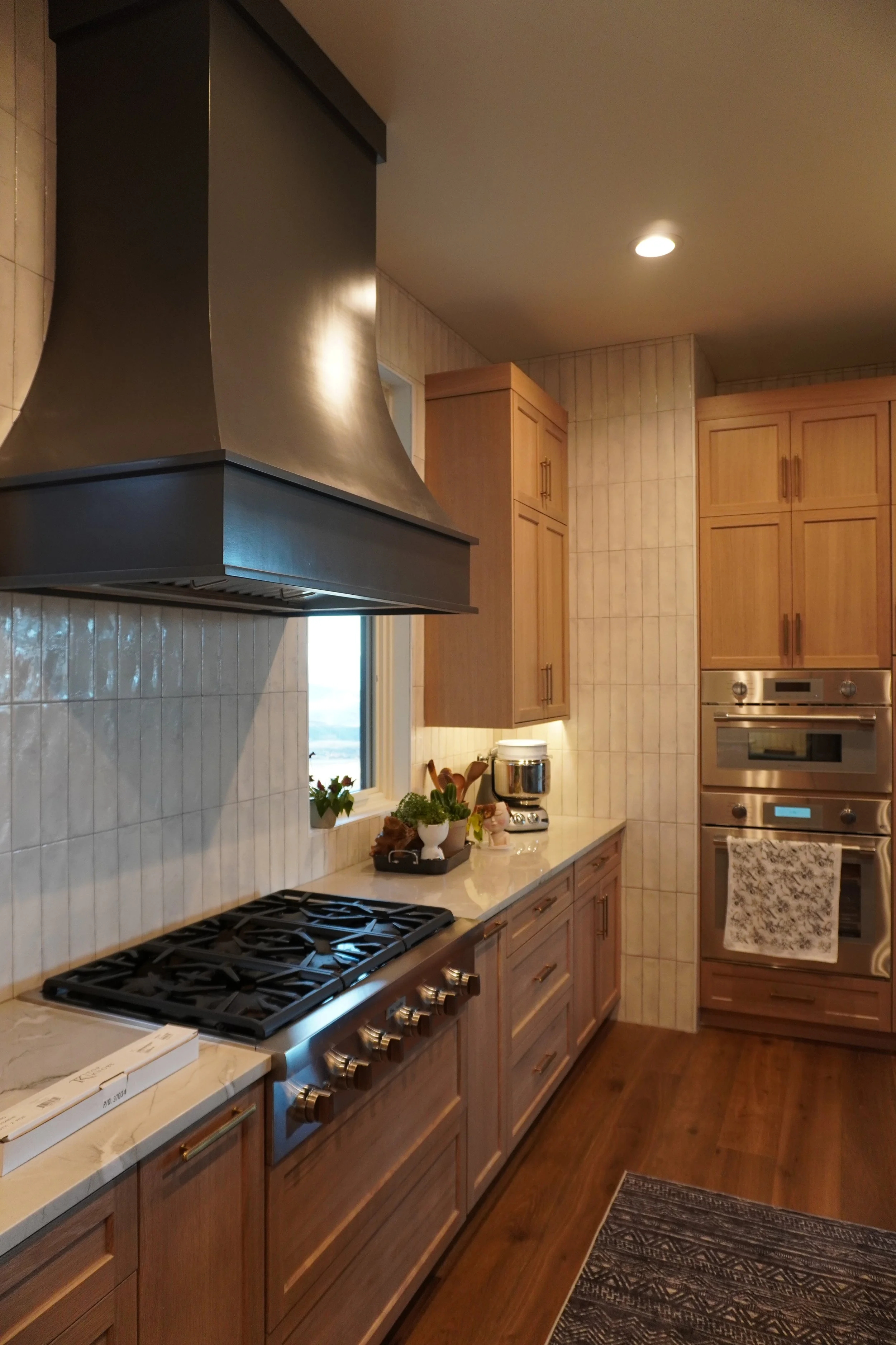 A kitchen with wooden cabinets, a stove with burner cooktop, a stainless steel range hood, a window, countertop appliances, and potted plants.