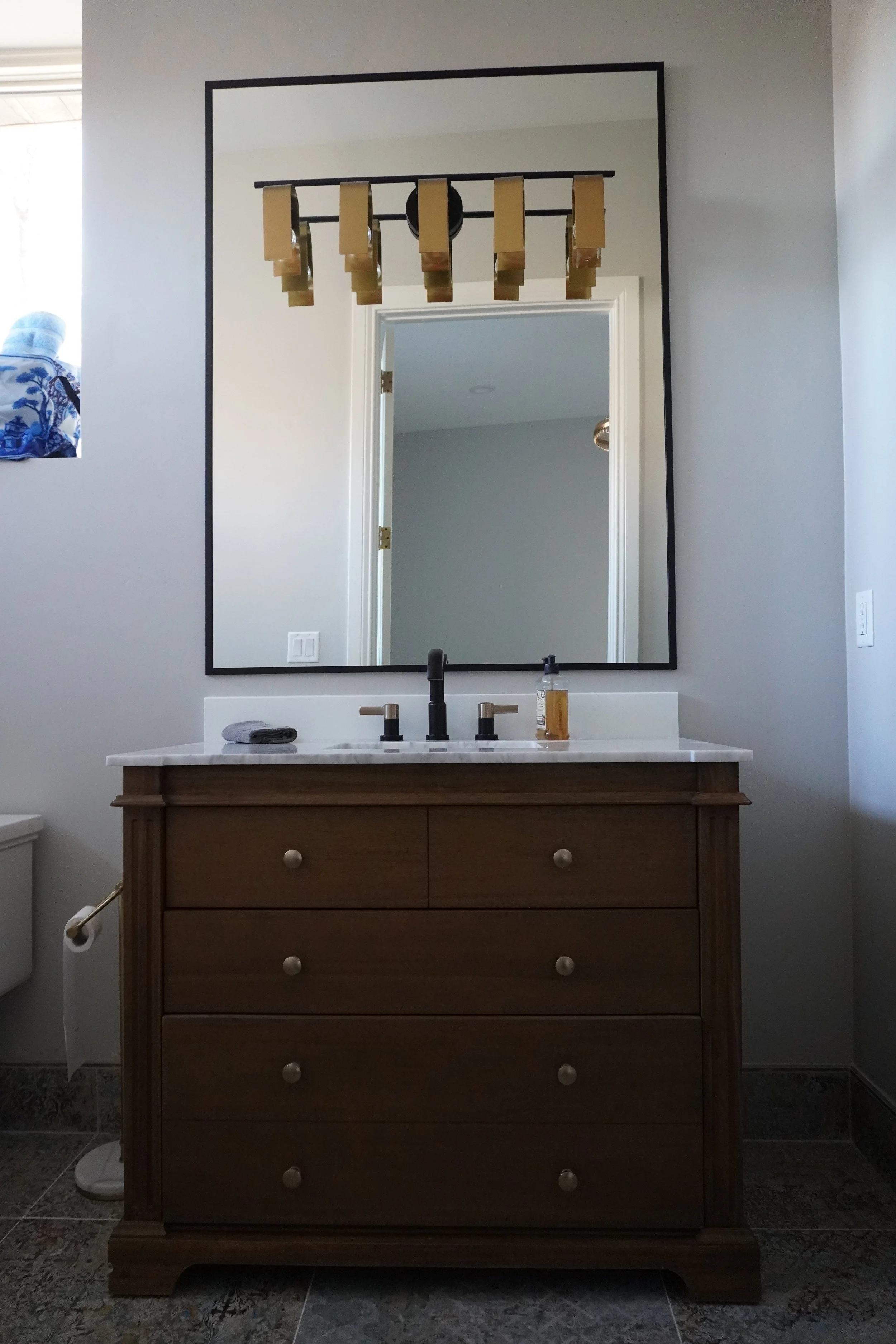 Bathroom with a wooden vanity, white countertop, and a large framed mirror. The mirror reflects a doorway and part of another room, with a lighting fixture resembling an industrial pipe chandelier across the top of the mirror.