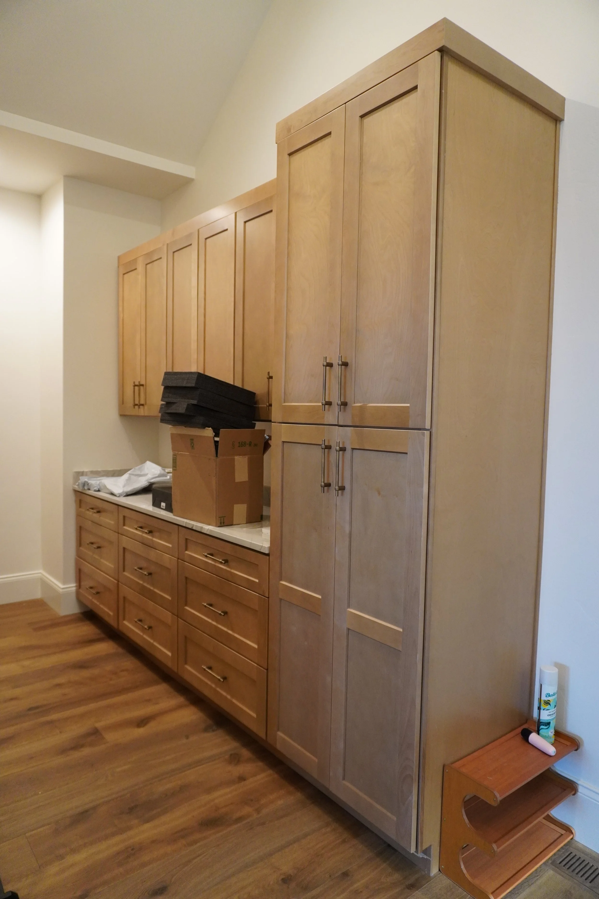 Wooden kitchen cabinets beside a countertop with a cardboard box and black foam pieces, a small wooden shelf with cleaning supplies, and a section of a hardwood floor.