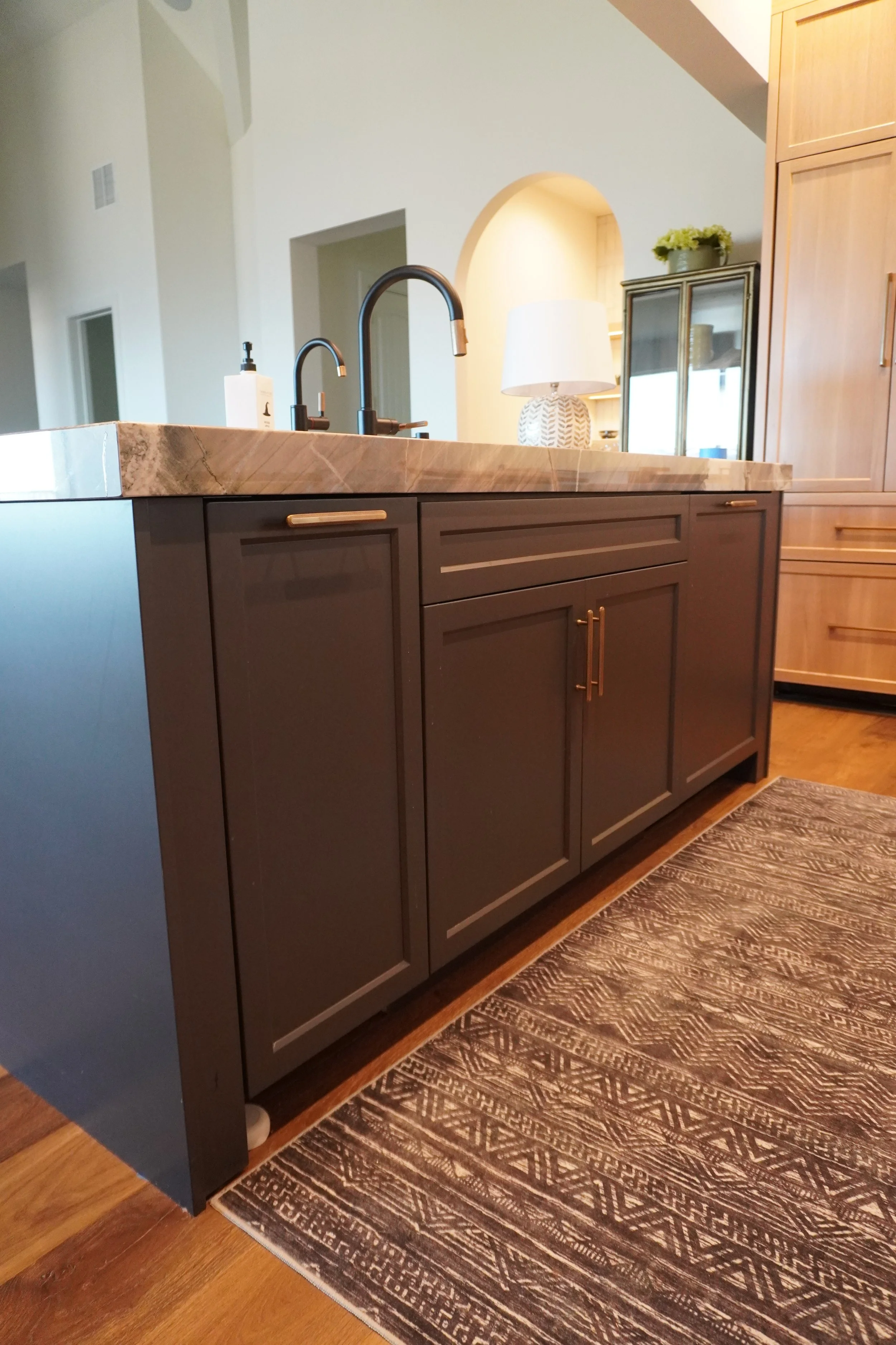 Kitchen island with dark cabinets, marble countertop, and dual sinks with black faucets. A patterned rug on wooden floor, and a lamp and cabinet in the background.