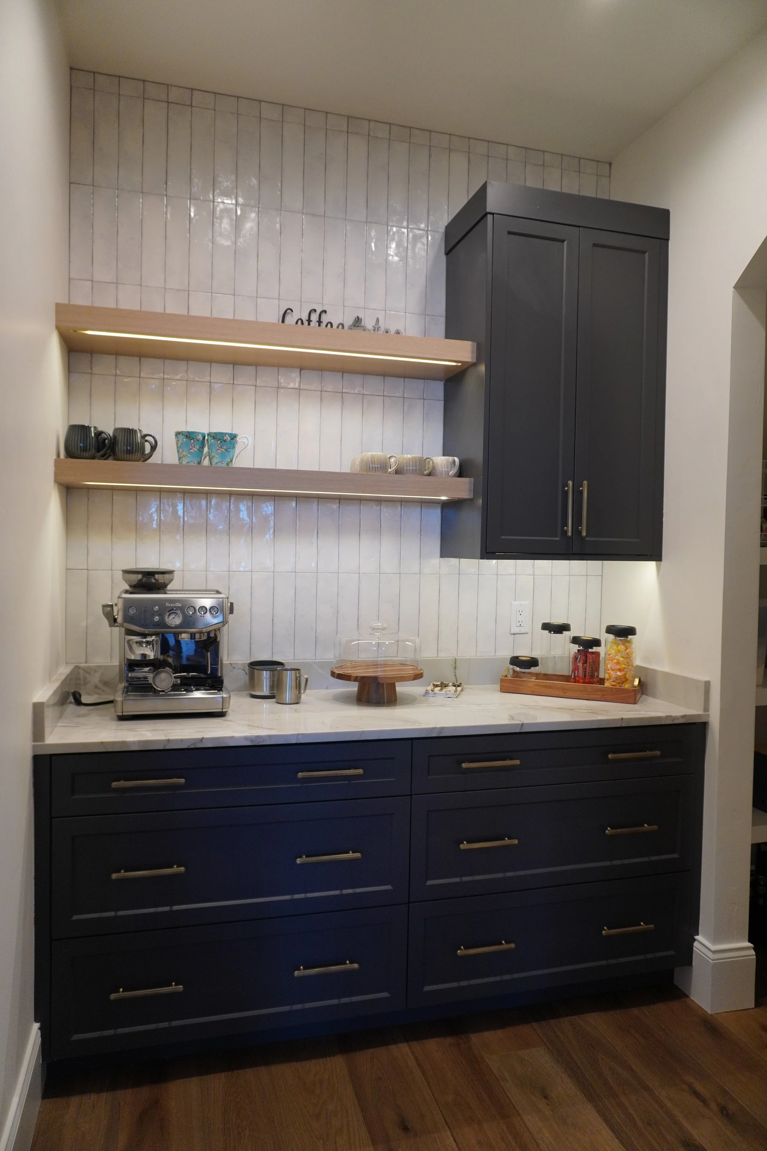 Kitchen corner with dark cabinetry, marble countertop, and open shelves with cups and mugs, coffee machine, cake stand, spice jars, and wall decor reading 'coffee & tea'. Default kitchen wall tiles.