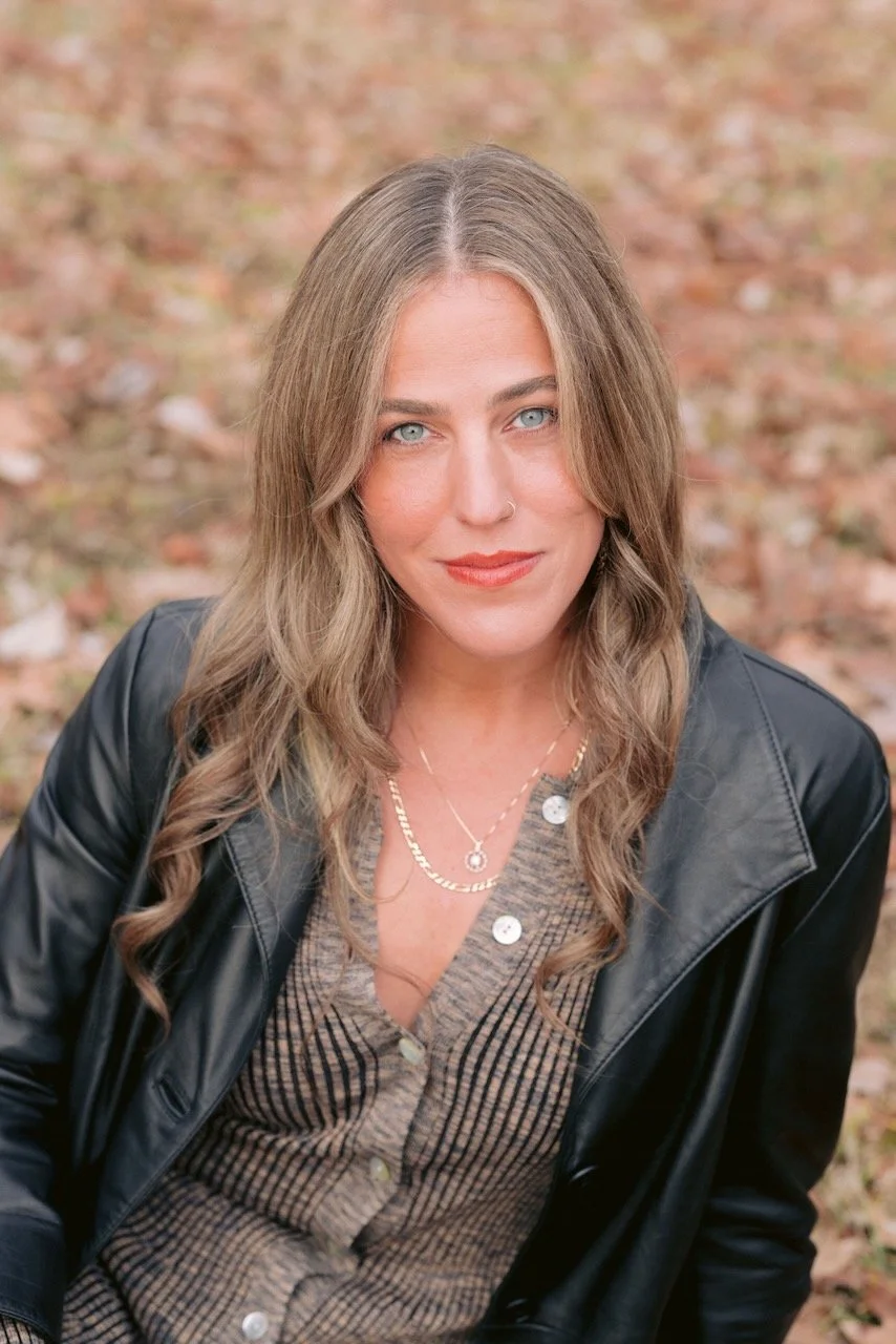 A woman with long wavy hair, blue eyes, and a nose piercing, wearing a checkered shirt, layered necklaces, and a black leather jacket, sitting outdoors on autumn leaves.