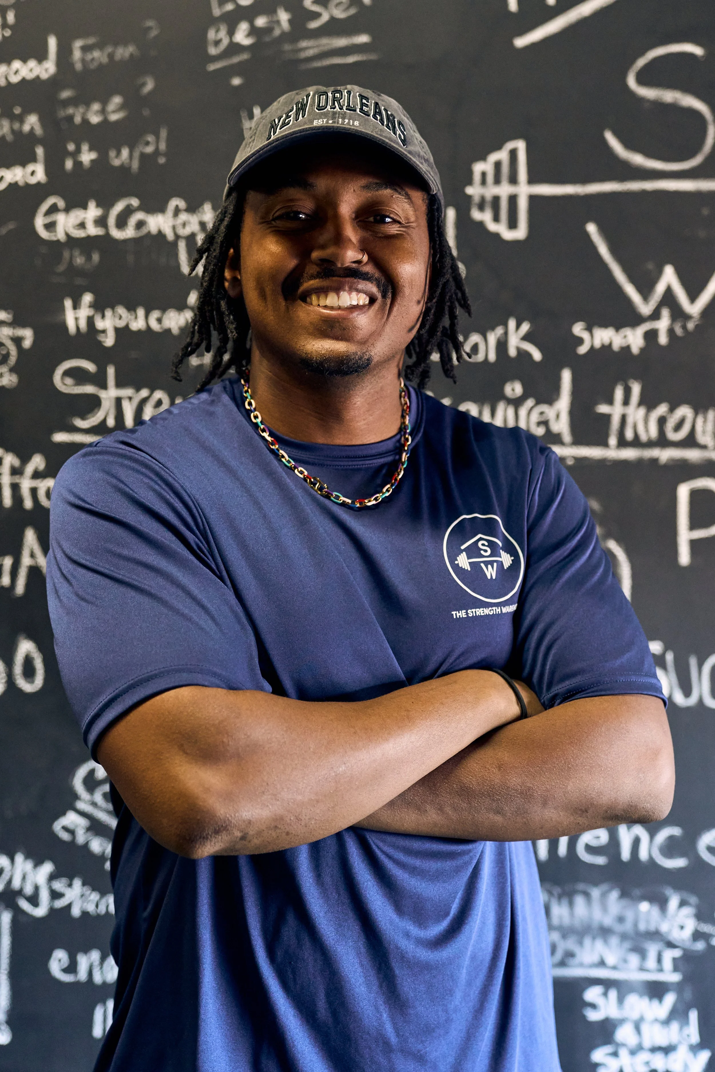 Mikael Ruffin, trainer, smiling and crossing his arms, wearing a navy blue t-shirt with The Strength Warehouse logo, standing in front of the Mindfulness Wall, full of clients' intentions for their workouts