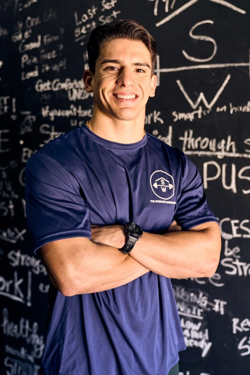 George Blevins, trainer, smiling and crossing his arms, wearing a navy blue t-shirt with The Strength Warehouse logo, standing in front of the Mindfulness Wall, full of clients' intentions for their workouts