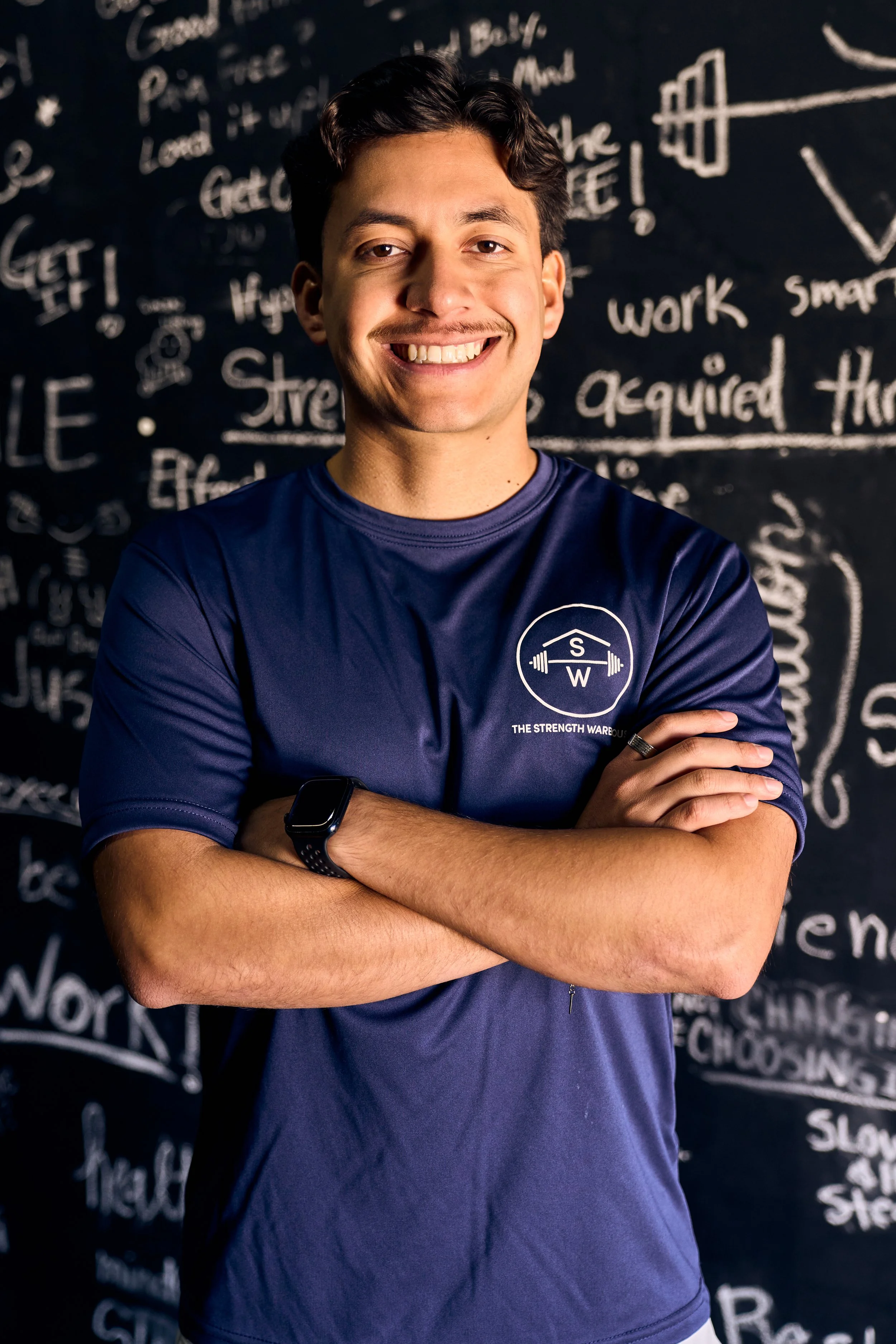 Adrian Jaimes, lead trainer and owner, smiling and crossing his arms, wearing a navy blue t-shirt with The Strength Warehouse logo, standing in front of the Mindfulness Wall, full of clients' intentions for their workouts